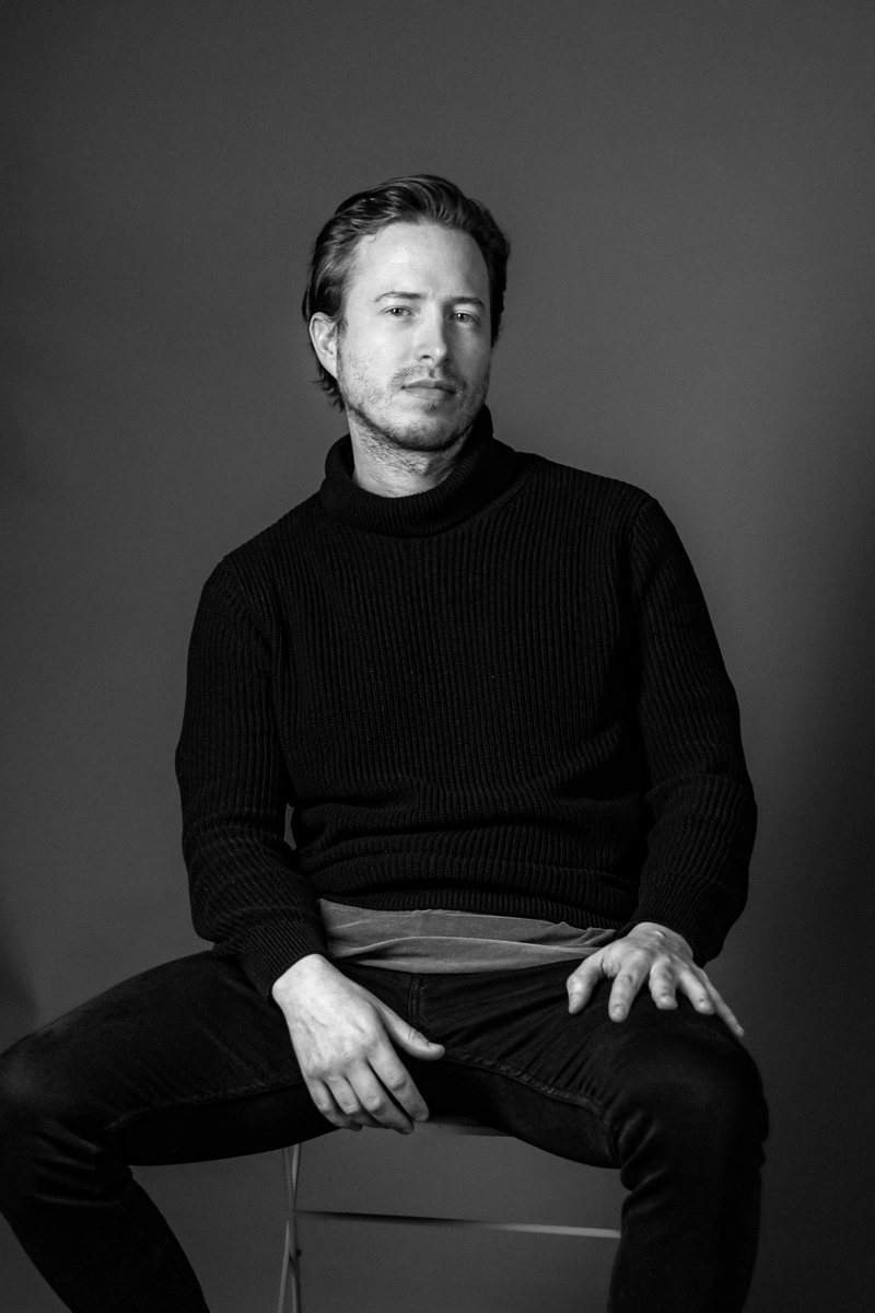 Black-and-white portrait of Alexander Main, seated, wearing a dark knit turtleneck